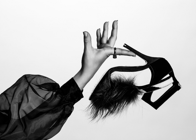  – Svartvitt fotografi av en högklackad sko med päls på tån, som hålls av en kvinna i en tunn puffblus