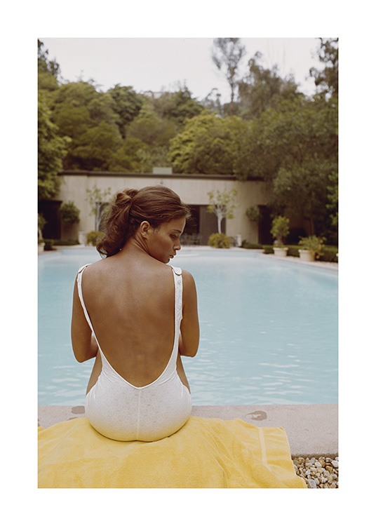  – Fotografi av en kvinna som sitter på en handduk framför en pool i en vit baddräkt