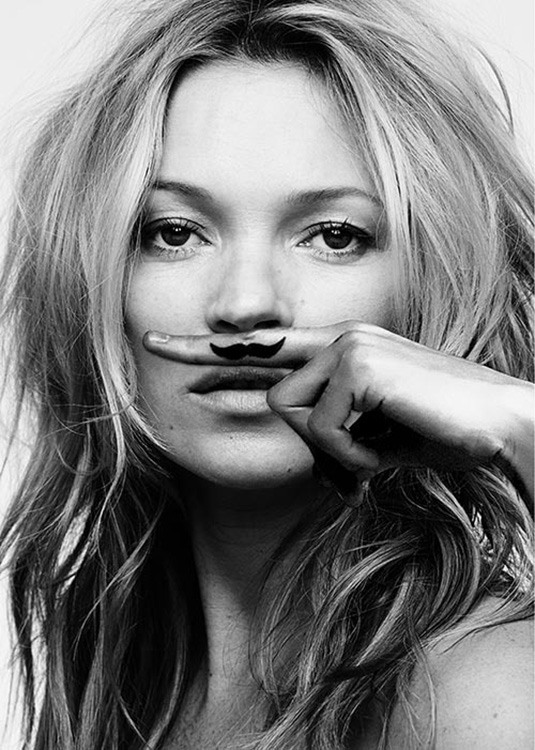  – Svartvitt fotografi av Kate Moss med ett finger ovanför munnen med en mustasch på fingret